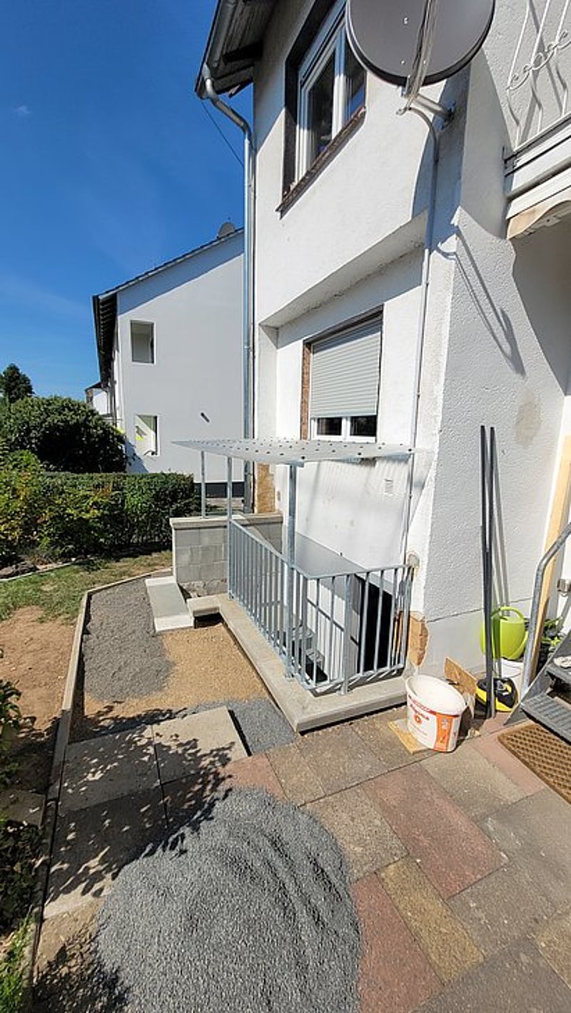Bau eines barrierefreien Zugangs mit Rampen und Geländer an einem Wohngebäude, umgeben von Kies und Garten.