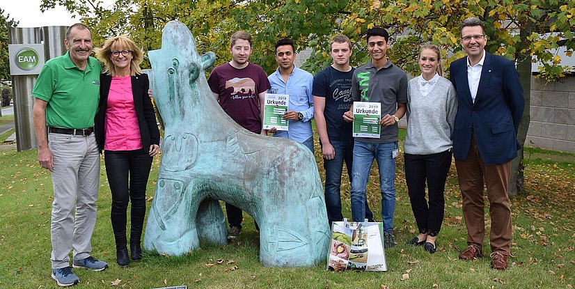 Gruppe von Menschen neben einer Skulptur, alle halten Urkunden in der Hand, im Hintergrund Bäume und ein Firmenlogo.
