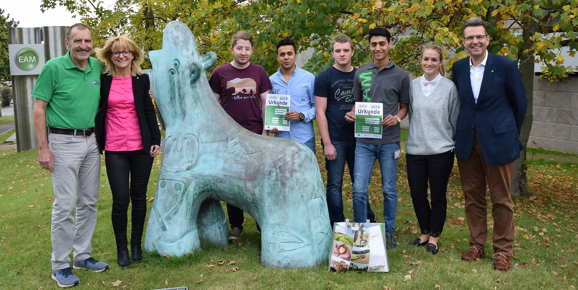 Gruppe von Menschen neben einer Skulptur, alle halten Urkunden in der Hand, im Hintergrund Bäume und ein Firmenlogo.