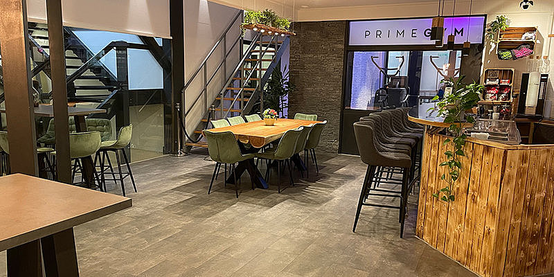 Moderne Cafélounge mit Holztisch, grünen Stühlen, Holztheke und Treppe, die in einen Fitnessbereich führt. Gute Beleuchtung.