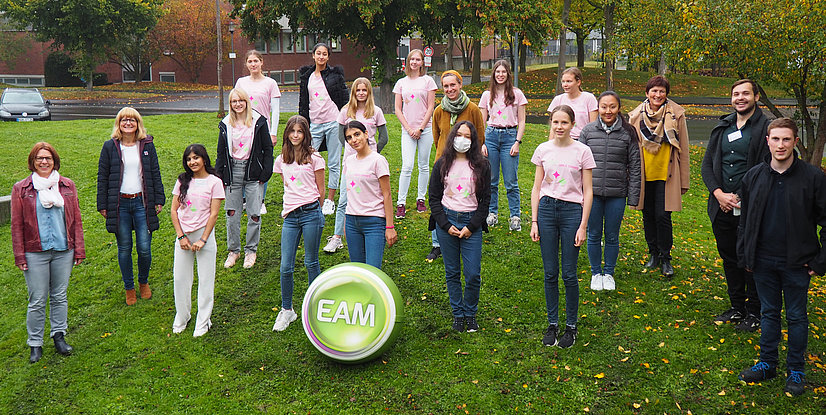 Gruppenfoto von Jugendlichen und Betreuern in pinken T-Shirts, um einen großen, grünen EAM-Ball, im Freien auf Rasen.