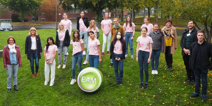 Gruppenfoto von Jugendlichen und Betreuern in pinken T-Shirts, um einen großen, grünen EAM-Ball, im Freien auf Rasen.