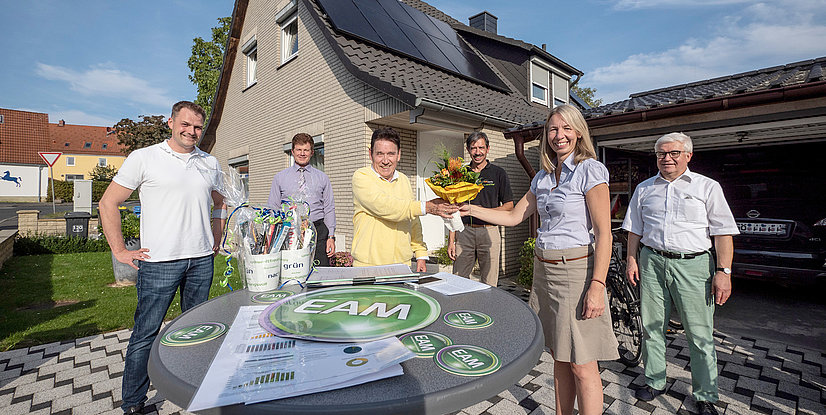 Sechs Personen stehen im Freien, während eine Person einen Blumenstrauß überreicht, vor einem Haus mit Solarpanelen.