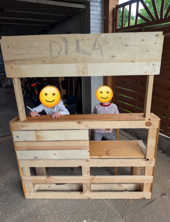 Zwei Kinder sitzen an einem selbstgebauten Holztisch mit einem Schild über ihnen. Eines schreibt, das andere schaut.