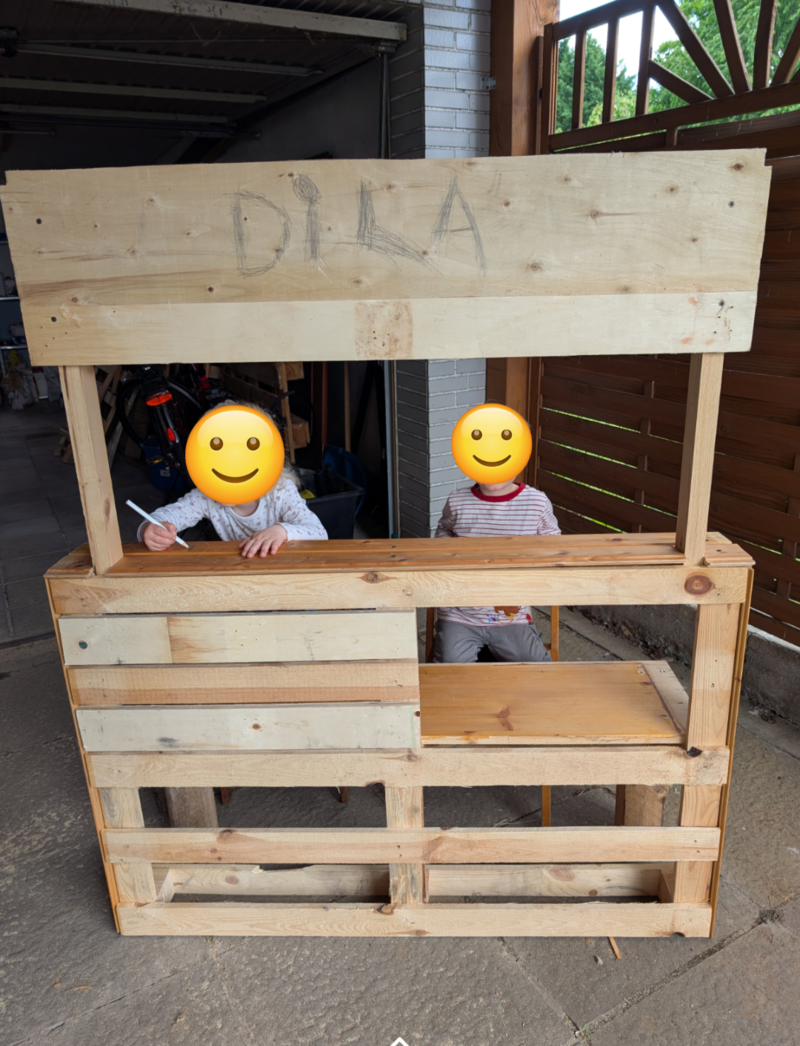 Zwei Kinder sitzen an einem selbstgebauten Holztisch mit einem Schild über ihnen. Eines schreibt, das andere schaut.
