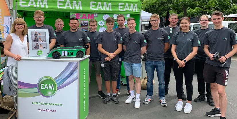  Eine Gruppe von jungen Menschen in grauen T-Shirts mit EAM-Logo stehen lächelnd neben einem grünen Modellrennwagen mit der Nummer 1. Im Hintergrund ist ein Banner mit EAM zu sehen.