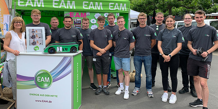 Eine Gruppe von jungen Menschen in grauen T-Shirts mit EAM-Logo stehen lächelnd neben einem grünen Modellrennwagen mit der Nummer 1. Im Hintergrund ist ein Banner mit EAM zu sehen.