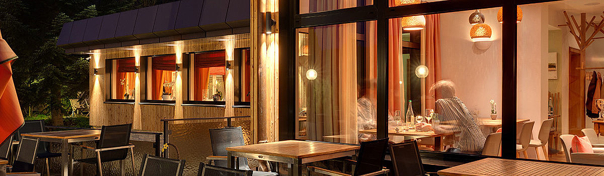 Angenehme Außenansicht eines Restaurants bei Nacht, Beleuchtung und Tischen zur Außensitzung sichtbar.