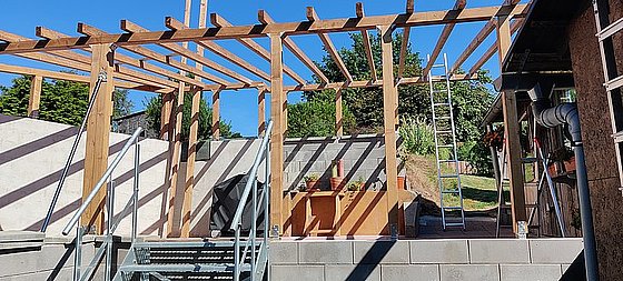 Baugerüst mit Holzstützen und Treppe, im Hintergrund Pflanzen und ein Kühlschrank, bei sonnigem Wetter.