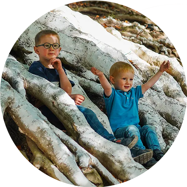 Zwei Kinder sitzen auf großen Wurzeln, der ältere schaut nachdenklich, der jüngere freut sich und hebt die Arme.