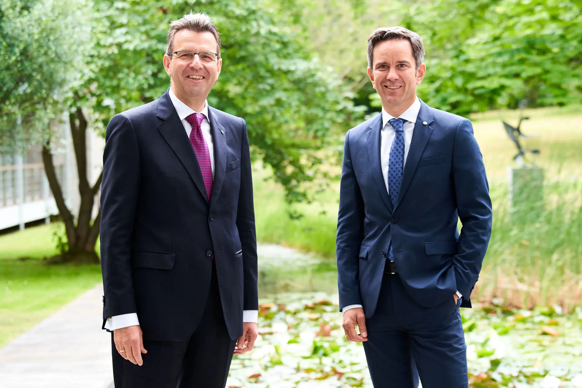 Zwei Männer in Anzügen stehen vor einer Grünfläche mit Teich und Pflanzen im Hintergrund.