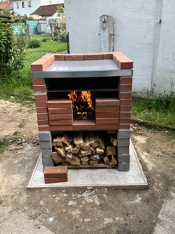 Gemauerter Grillofen mit loderndem Feuer und Lagerholz, umgeben von grüner Wiese und Innenhof.