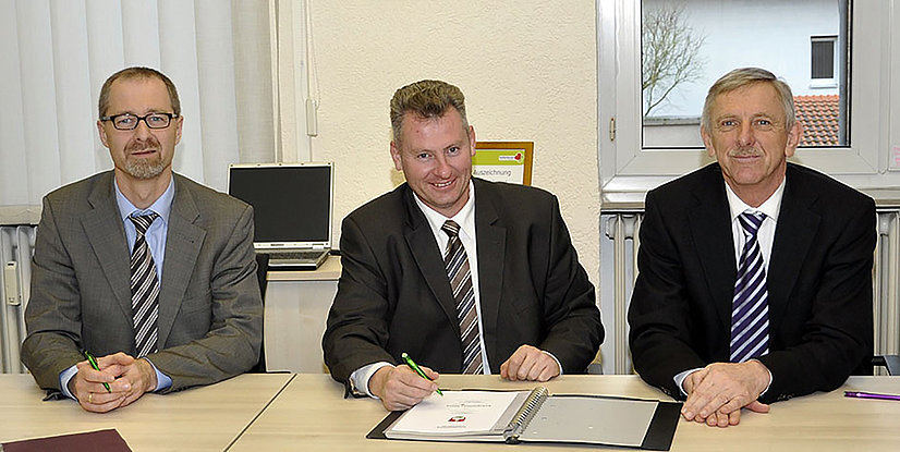 Drei Männer in Anzügen sitzen an einem Tisch mit Dokumenten und einem Laptop im Hintergrund.
