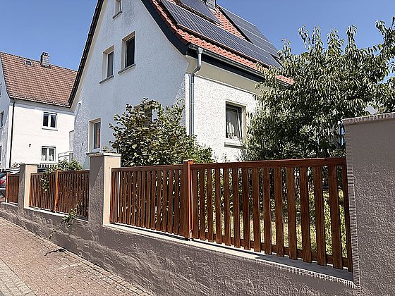 Weißes Wohnhaus mit rotem Dach, Solarpanelen, umgeben von grüner Vegetation und einem Holzzaun.