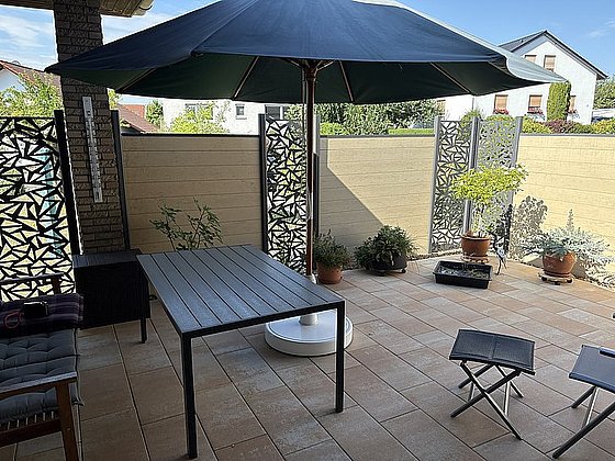 Gemütlicher Patio mit einem Tisch, Stühlen, einem großen Sonnenschirm und dekorativen Pflanzen. Helle Fliesenboden und Blumen.