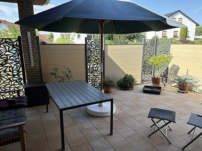 Gemütlicher Patio mit einem Tisch, Stühlen, einem großen Sonnenschirm und dekorativen Pflanzen. Helle Fliesenboden und Blumen.