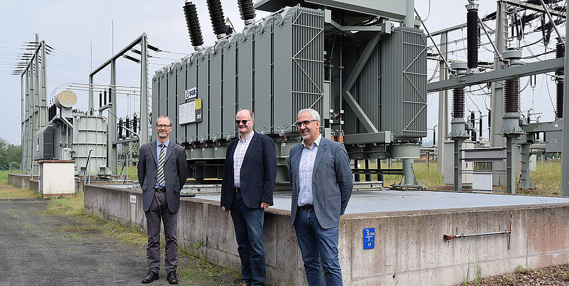 Drei Personen stehen vor einem Transformator in einer Energieanlage, umgeben von elektrischen Geräten und Hochspannungsmasten.