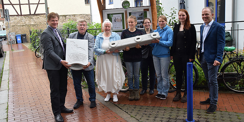 Gruppierung von Personen, die eine Skulptur oder ein Kunstwerk halten, in einer urbanen Umgebung mit Fahrrädern im Hintergrund.