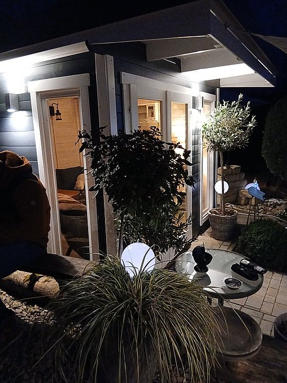 Gemütliche Veranda bei Nacht mit Beleuchtung, Pflanzen und Glastisch, ideal für Entspannung im Freien.