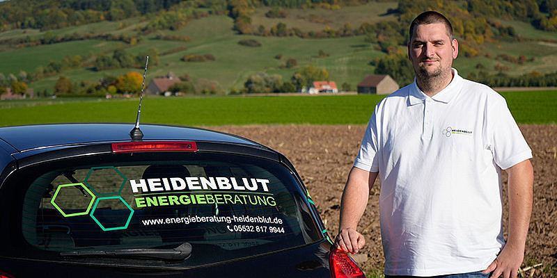 Mann steht neben einem Auto mit Werbung für Heidenblut Energieberatung, im Hintergrund eine Landschaft mit Bäumen.