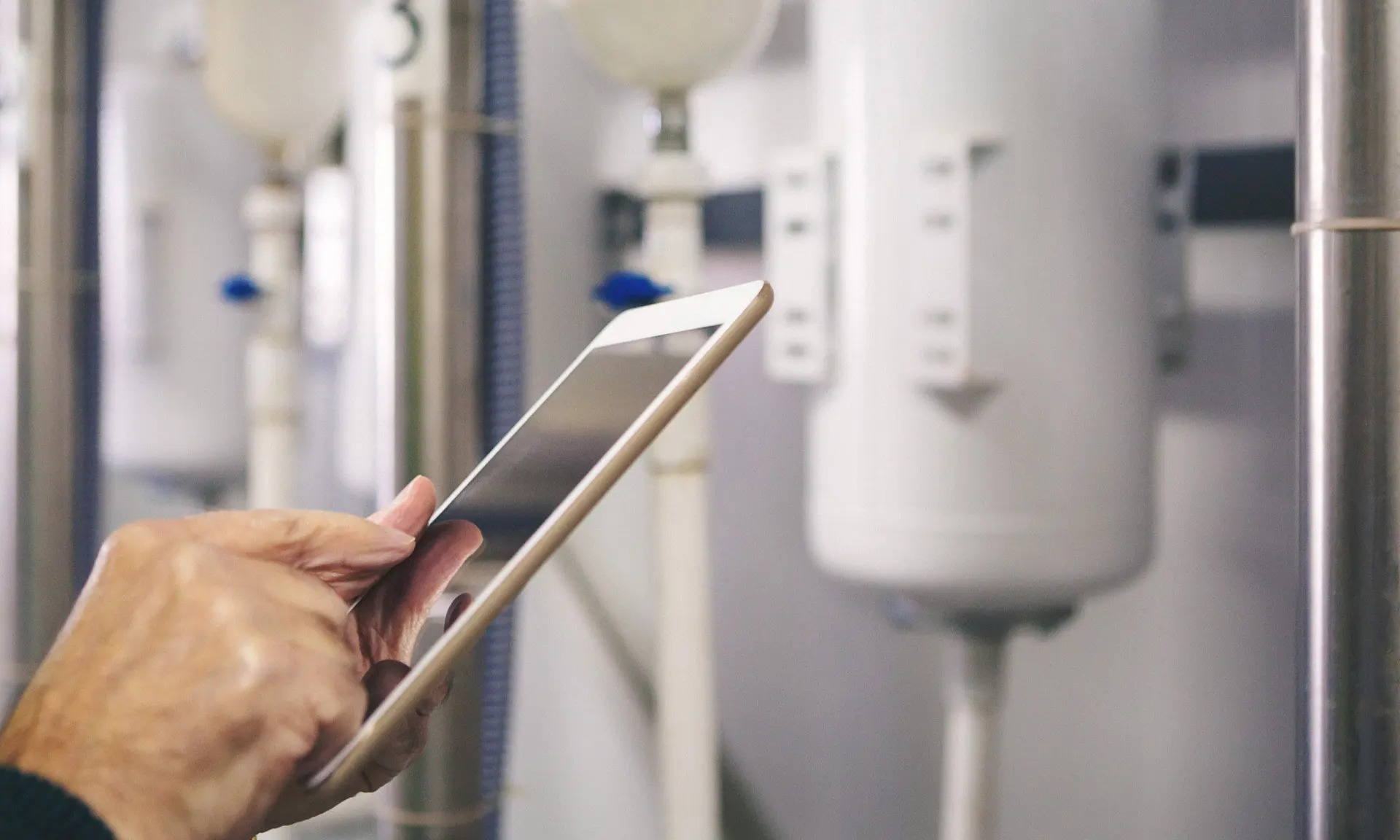 Eine Hand hält ein Smartphone schräg vor einem weißen Hintergrund mit technischen Geräten und Rohren.