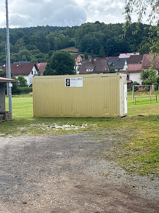 Gelber Container auf einer Wiese, umgeben von Bäumen und Häusern im Hintergrund, mit Schildern und Gras.
