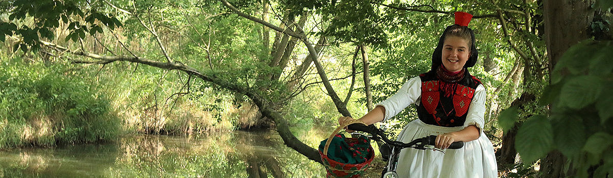 Mädchen in traditioneller Tracht mit Fahrrad am Ufer eines ruhigen Gewässers, umgeben von Bäumen und Natur.