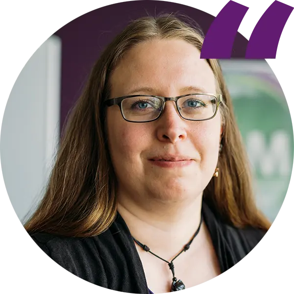 Porträt einer Person mit langen Haaren und Brille, lächelnd, vor lila Hintergrund, Quote-Symbol oben rechts.