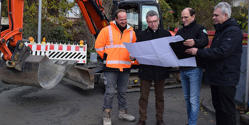 Vier Personen in Arbeitskleidung besprechen Pläne vor einer Baumaschine auf einer Baustelle.
