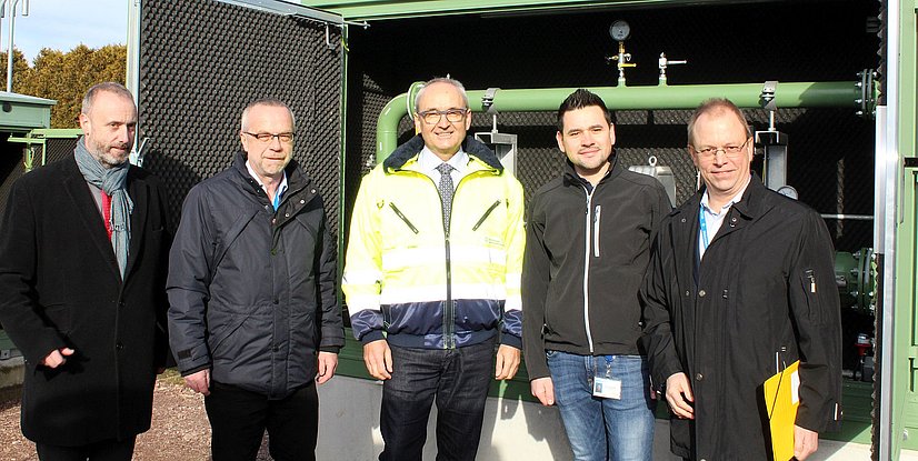 Gruppe von fünf Männern steht vor technischen Geräten im Freien, alle tragen Arbeitskleidung und schauen zur Kamera.