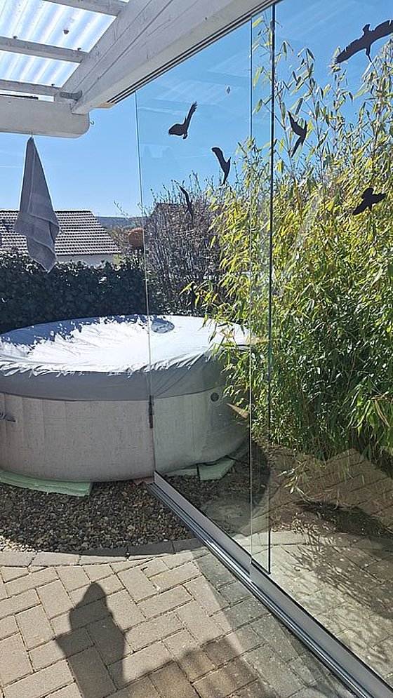 Ein runder Whirlpool mit Abdeckung in einem sonnigen Garten, umgeben von Bambus und einer Glaswand.
