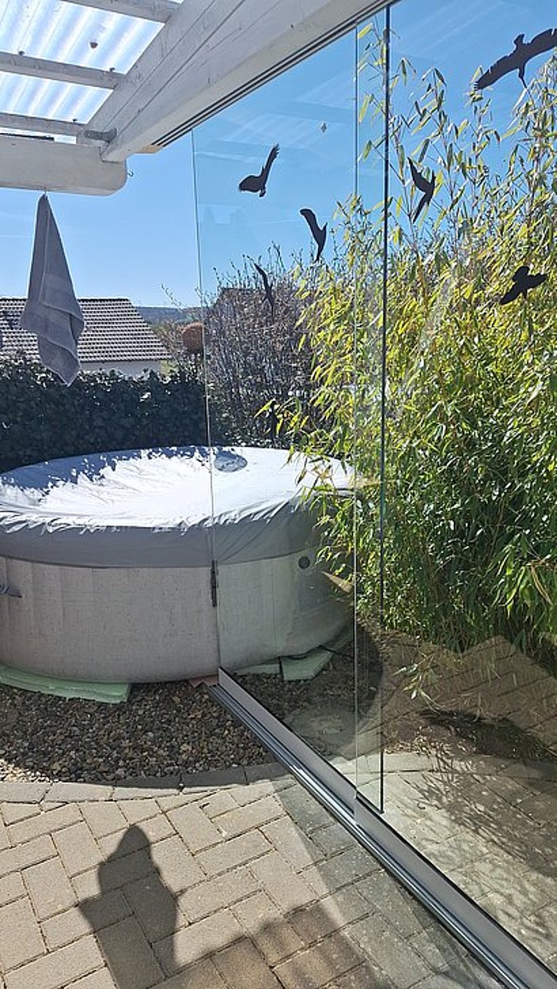 Ein runder Whirlpool mit Abdeckung in einem sonnigen Garten, umgeben von Bambus und einer Glaswand.