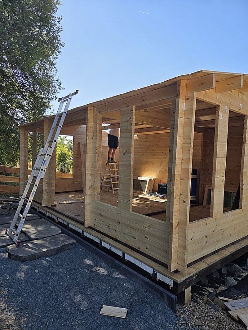 Bau eines Holzhauses mit Leiter und Handwerker, der an den Wänden arbeitet, bei klarem Himmel.