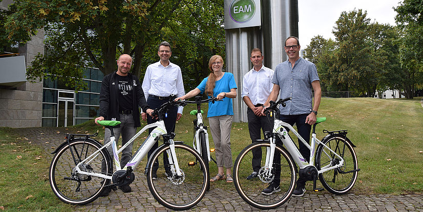 Fünf Personen stehen lächelnd neben ihren E-Bikes vor einem Firmengebäude mit grünem Logo im Freien.