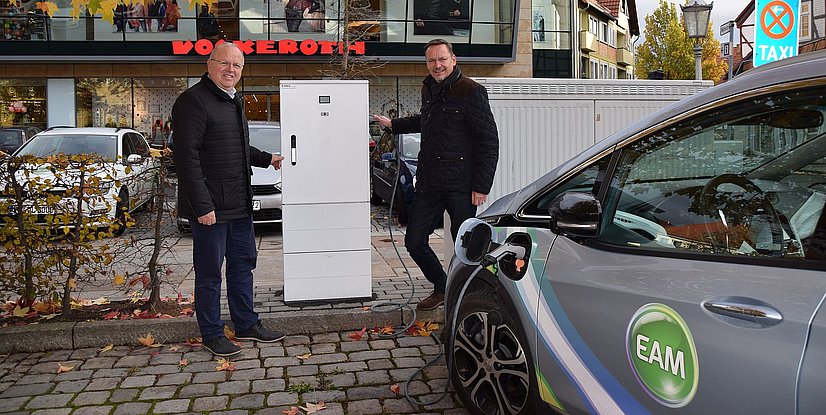 Zwei Männer stehen neben einer Ladestation für Elektroautos, im Hintergrund ist ein Geschäft zu sehen. Herbstliche Umgebung.