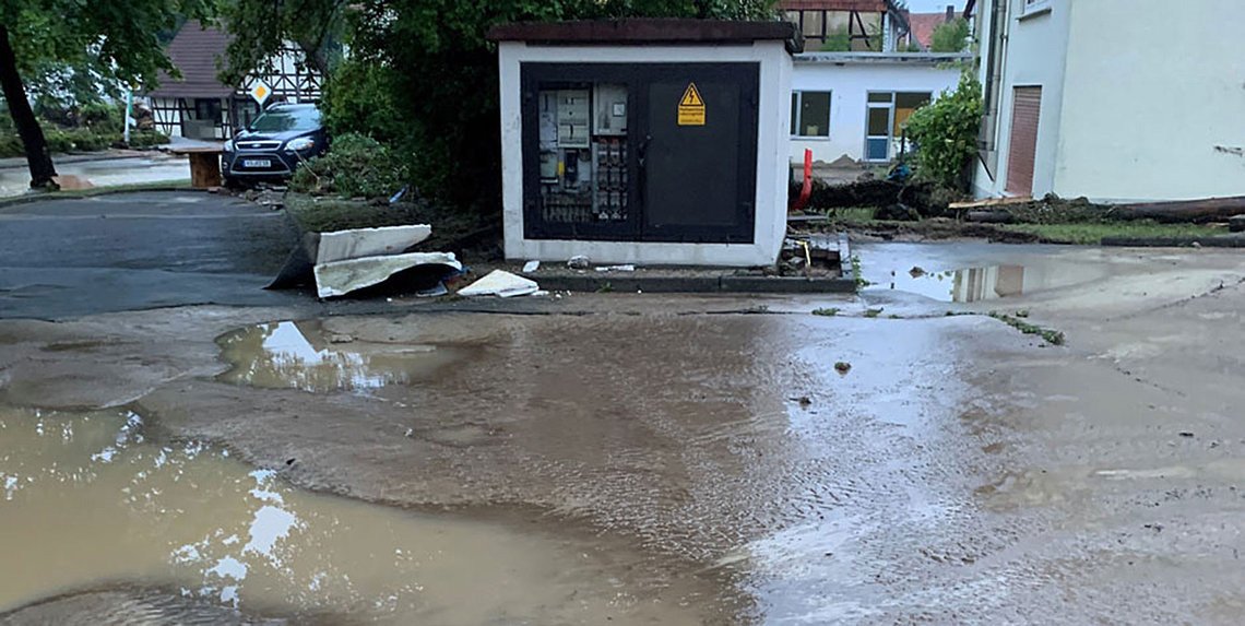 Eine schwarze Trafostation mit gelbem Warnschild steht inmitten einer mit Schlamm bedeckten Straße mit Pfützen und Trümmern nach einem Unwetter.