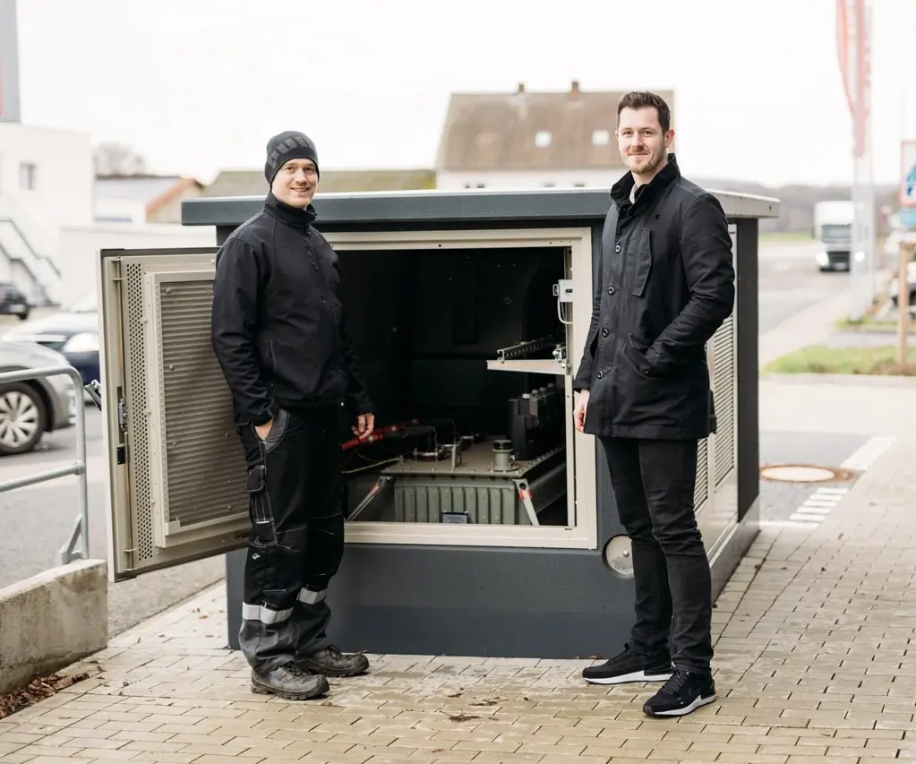 Zwei Männer stehen vor einem geöffneten technischen Schrank auf der Straße, umgeben von urbaner Umgebung.