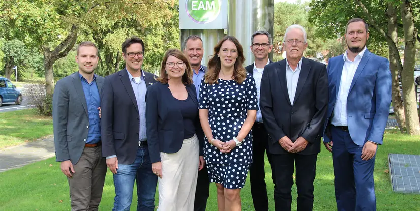 Gruppenbild vor einem EAM-Schild, umgeben von Bäumen und Rasen. Alle lächeln und stehen auf einem grünen Grundstück.