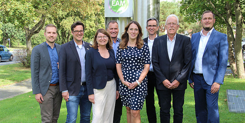 Gruppenbild vor einem EAM-Schild, umgeben von Bäumen und Rasen. Alle lächeln und stehen auf einem grünen Grundstück.