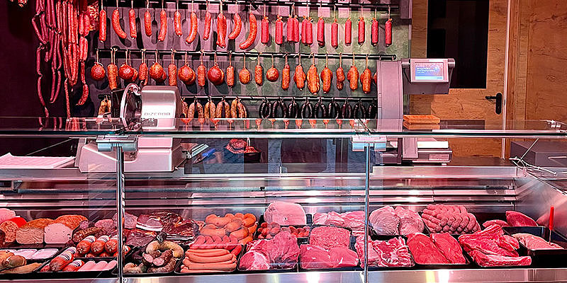 Metzgerei mit einer Auswahl an Wurst- und Fleischwaren, präsentiert in einer gläsernen Theke und an der Wand hängend.