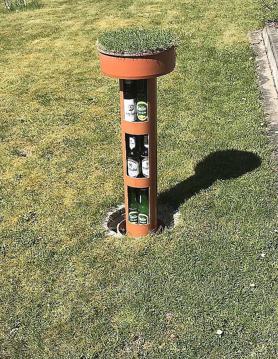 Ein Pflanzgefäß in Form eines Standrohrs mit Gras und seitlichen Flaschenablagen auf einem Rasenfläche.