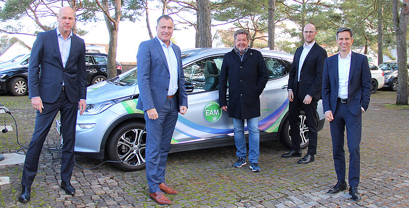 Fünf Personen in Anzügen stehen neben einem Elektroauto auf einem Parkplatz, umgeben von Bäumen und anderen Fahrzeugen.