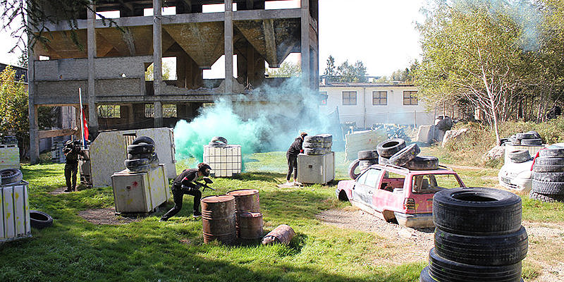 Zwei Spieler in einem Paintballfeld, umgeben von Autowracks und verdeckenden Barrikaden, während grüner Rauch sichtbar ist.