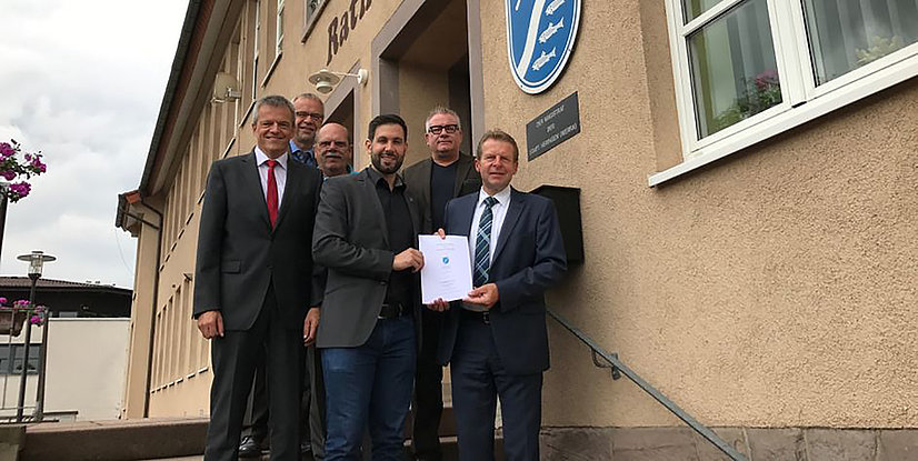 Sechs Personen posieren vor einem Gebäude, während einer einen Dokumentenübergabe durchführt.