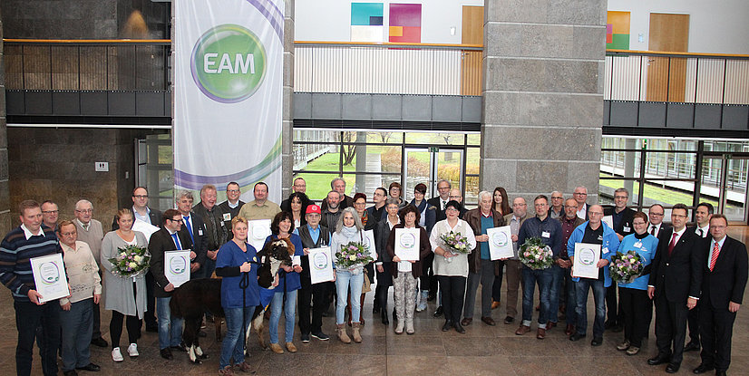 Gruppenfoto von Personen mit Blumen und Urkunden in einem modernen Gebäude, EAM-Logo im Hintergrund sichtbar.