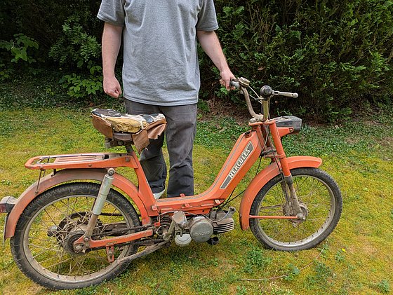 Mann mit grauem T-Shirt steht neben einem orangefarbenen Moped mit abgenutztem Sattel in einem grünen Garten.