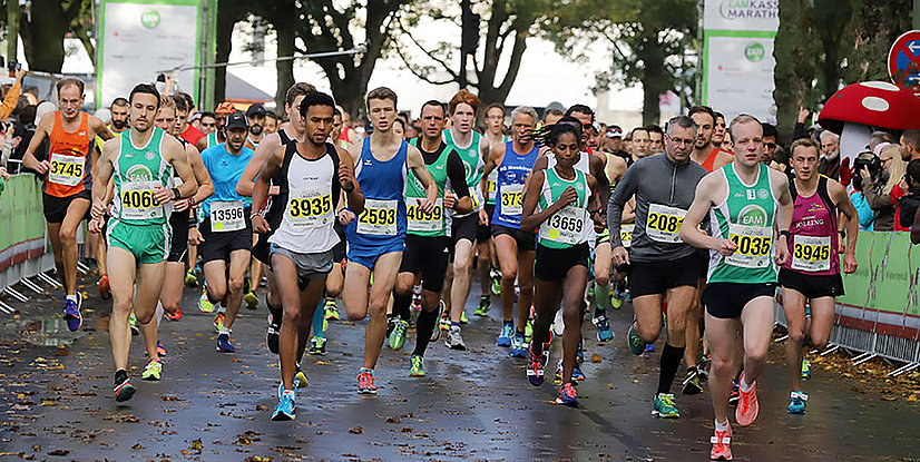 Eine Gruppe von Läufern beim Start eines Marathons auf einer Asphaltstraße, umgeben von Zuschauern und Werbebannern.