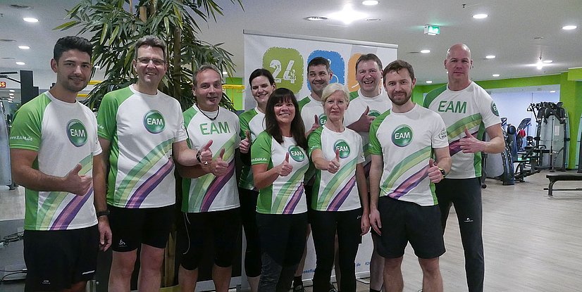 Vierzehn Personen in Sportkleidung mit EAM-Logo posieren in einem Fitnessstudio vor einem Banner mit der Aufschrift 24 Stunden