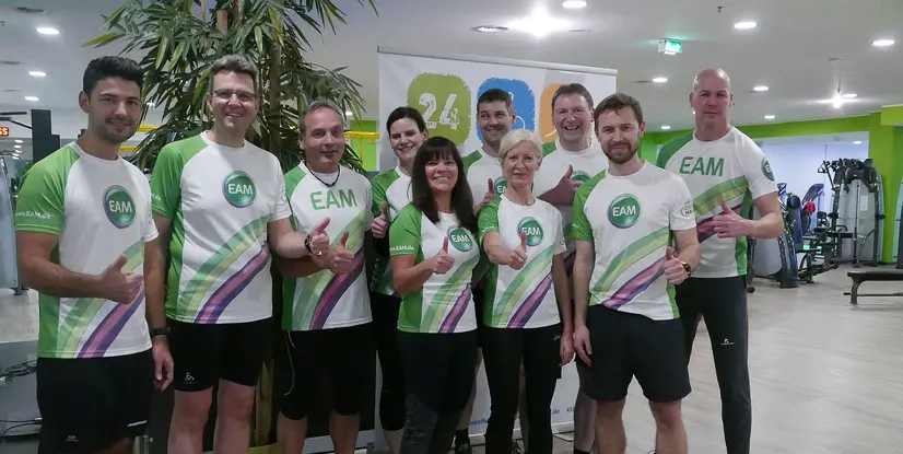 Vierzehn Personen in Sportkleidung mit EAM-Logo posieren in einem Fitnessstudio vor einem Banner mit der Aufschrift 24 Stunden