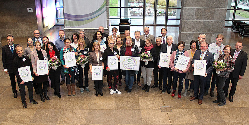 Gruppenfoto von Menschen mit Urkunden und Blumen in einem modernen Raum, umgeben von Fenster und Steinwand.